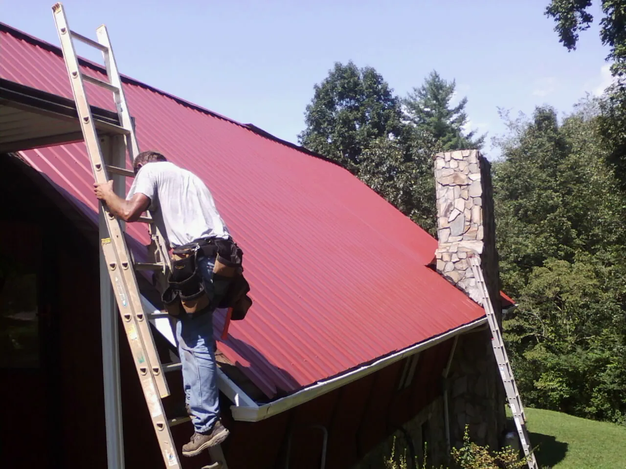 Roofer on a ladder beside a red metal roof and stone chimney — Soffit & Fascia Repair by Roofing Brothers in North Fairfield